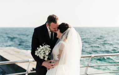 Güzel gelin ve damat, iskelede denizin arka planında sarılıyorlar. Denizde güneşli bir yaz gününde düğün günü. Deniz kenarında bir çiftin yürüyüşü. Mutlu evlilik çifti