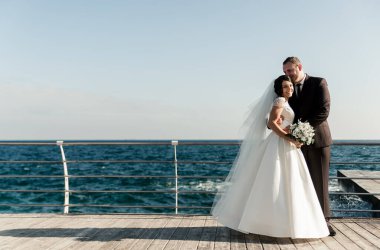 Güzel gelin ve damat, iskelede denizin arka planında sarılıyorlar. Denizde güneşli bir yaz gününde düğün günü. Deniz kenarında bir çiftin yürüyüşü. Mutlu evlilik çifti