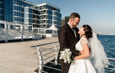 Güzel gelin ve damat, iskelede denizin arka planında sarılıyorlar. Denizde güneşli bir yaz gününde düğün günü. Deniz kenarında bir çiftin yürüyüşü. Mutlu evlilik çifti