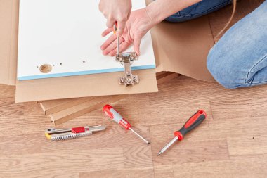 Bir insan mobilya montajı yaparken araçlar yerde düzenlenir. Dekor ahşap döşeme ve geniş doğal ışık içeriyor. Detaylara dikkat etmek işinizde barizdir.