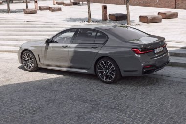 BMW 740D. Lüks bir sedan, taştan basamaklar ve modern banklarla çevrili, zarif vücudunu ve gelişmiş tasarımını gün ışığında sergileyen asfaltlı bir caddeye park eder. Katowice, Polonya, 13.06.2021