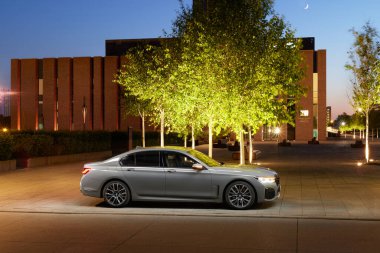 BMW 740D iyi aydınlatılmış bir patikanın yanında park halinde. Ağaçları çevreleyen ve arabanın zarif tasarımını geliştiren NOSPR konser salonunun modern binası. Katowice, Polonya, 13.06.2021