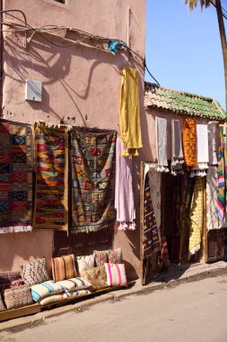 Parlak halılar ve renkli yastıklar bir mağazanın önünde satışa sunuluyor. Burası Fas, Marakeş Medine 'de gündüz vakti işlek bir alışveriş caddesi.
