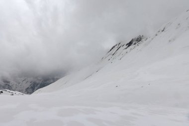 Uzakta ağır bulutlarla örtülü kayalık dağlarla birlikte engin bir kar tarlası sahneyi kaplıyor. Nepal 'deki Annapurna Ana Kampı yolunda.
