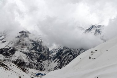 Gezginler, Annapurna Ana Kampı 'na giden Himalayalar' ın karlı arazisinde geziniyorlar. Yüksek dağ zirveleri kalın ve alçak bulutlar tarafından kısmen gizleniyor..