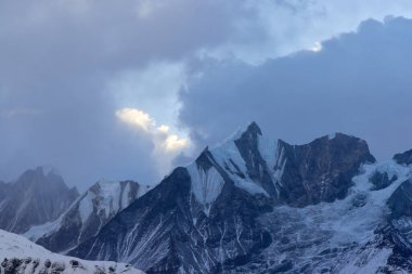 Karla kaplı dramatik dağ zirveleri Nepal 'deki Annapurna Ana Kampı' nda görülebilir. Bulutlar gökyüzünü dolduruyor. Muhteşem Himalaya manzarası üzerinde kasvetli bir atmosfer yaratıyor..