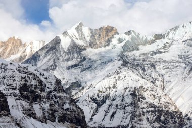 Karlı dağlar, kaya oluşumları ve buzullar parçalı bulutlu bir gökyüzünün altında duruyor. Manzara Nepal Himalayalardaki Annapurna Sığınağı 'nın içinden..