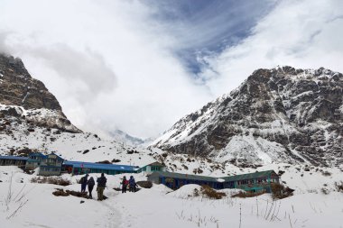 Bir grup yürüyüşçü karlı bir yolda bir kulübeye yaklaşıyor. Nepal 'deki Annapurna Ana Kampı' na giderken bulutlu gökyüzü ile arka planda dağlar yükselir..