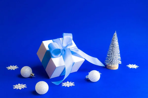 Świąteczne tło. Białe pudełko z niebieską wstążką, drzewo zimowe, płatki śniegu i srebrne kulki w składzie xmas na niebieskim tle dla kartki okolicznościowej. Dekoracja i kopiowanie miejsca na tekst. — Zdjęcie stockowe