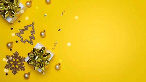 Surprise gift. White gift box with golden ribbon, New Year balls and sparkling lights in Christmas composition on dark yellow background for greeting card. Flat lay, top view, copy space.