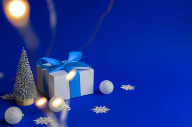 Noel arka planı mavisi. Mavi kurdeleli beyaz hediye kutusu, kış ağacı, Xmas kompozisyonunda kar taneleri ve gümüş toplar tebrik kartı için mavi arka planda. Metnin için dekorasyon ve kopyalama alanı