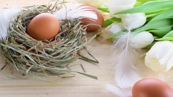 Paaseieren. Happy Easter decoratie: natuurlijke kleur eieren in mand met lente tulpen, witte veren op houten tafel achtergrond. Traditionele decoratie in zonlicht. Bovenaanzicht. — Stockvideo