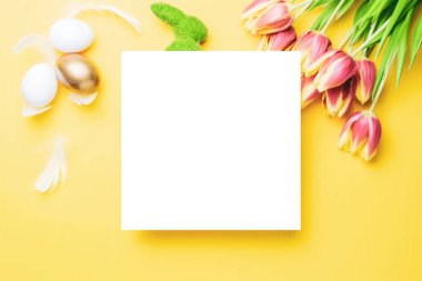 Paskalya mesajı. Bahar laleleriyle sepetin içinde altın, beyaz yumurta, Mutlu Paskalya dekorasyonunda pastel sarısı arka planda tüyler. Folyo minimalist yumurta tasarımı, modern üst görünüm şablonu