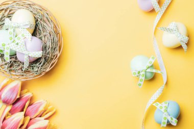 Yumurta rengi. Mutlu Paskalya süslemeleri: bant kurdeleli renkli yumurta, bahar laleleri, pastel sarısı arka planda beyaz tüyler. Folyo minimalist yumurta tasarımı, modern üst görünüm şablonu