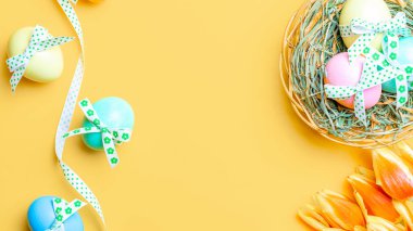 Yumurta rengi. Mutlu Paskalya süslemeleri: bant kurdeleli renkli yumurta, bahar laleleri, pastel sarısı arka planda beyaz tüyler. Folyo minimalist yumurta tasarımı, modern üst görünüm şablonu