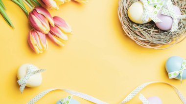 Yumurta rengi. Mutlu Paskalya süslemeleri: bant kurdeleli renkli yumurta, bahar laleleri, pastel sarısı arka planda beyaz tüyler. Folyo minimalist yumurta tasarımı, modern üst görünüm şablonu