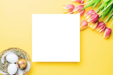 Paskalya seti. Bahar laleleriyle sepetin içinde altın, beyaz yumurta, Mutlu Paskalya dekorasyonunda pastel sarısı arka planda tüyler. Kopyalama alanı olan geleneksel tasarım