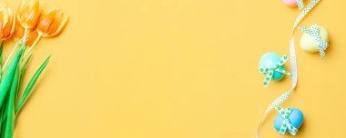 Paskalya geçmişi. Şerit kurdeleli renkli yumurta, bahar laleleri, Mutlu Paskalya dekorasyonunda pastel sarısı arka planda tüyler. Bahar tatili afişi