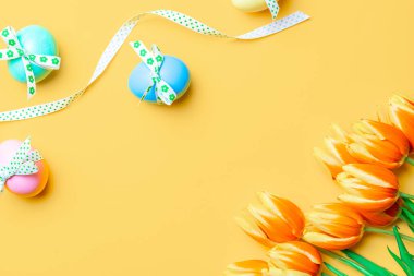 Yumurta rengi. Mutlu Paskalya süslemeleri: bant kurdeleli renkli yumurta, bahar laleleri, pastel sarısı arka planda beyaz tüyler. Folyo minimalist yumurta tasarımı, modern üst görünüm şablonu
