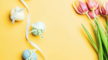 Paskalya tavşanı. Şerit kurdeleli renkli yumurta, bahar laleleri, Mutlu Paskalya dekorasyonunda pastel sarısı arka planda tüyler. Üst görünümde geleneksel tasarım