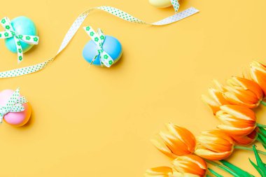 Yumurta rengi. Mutlu Paskalya süslemeleri: bant kurdeleli renkli yumurta, bahar laleleri, pastel sarısı arka planda beyaz tüyler. Folyo minimalist yumurta tasarımı, modern üst görünüm şablonu