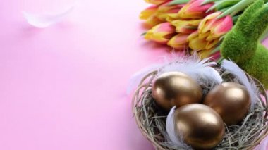 Paskalya yumurtaları sepetin içinde altın renginde bahar laleleri, pastel pembe arka planda beyaz tüyler Mutlu Paskalya süslemeleri. Şenlikli dekorasyon