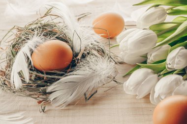 Sepette bahar laleleri olan renkli yumurtalar, Mutlu Paskalya dekorasyonunda ahşap masa arkasında beyaz tüyler. Folyo minimalist yumurta tasarımı, modern tasarım şablonu