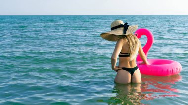 Sahilde bir kadın. Hasır şapkalı, bikinili, mavi okyanus suyunda pembe şişirilebilir flamingolu güneş gözlüklü seksi bir kadın. Seyahat tatili. Güzellik, sağlık, yaşam tarzı.