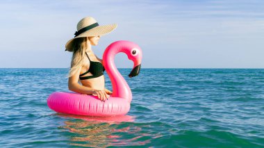 Genç kadınlar plajı. Hasır şapkalı, bikinili, mavi okyanus suyunda deniz arkası için şişirilebilir pembe flamingolu güneş gözlüklü seksi bir kadın. Yaz tatili eğlence özgürlüğü kavramı