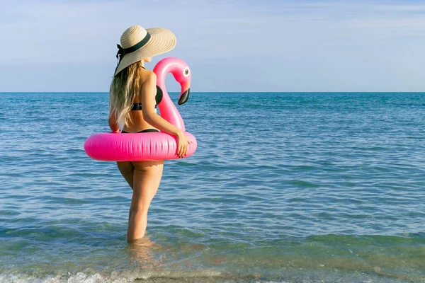 Yaz zamanı. Hasır şapkalı, bikinili, mavi okyanus suyunda pembe şişirilebilir flamingolu güneş gözlüklü seksi bir kadın. Yüzme suyu, eğlenme ve seyahatin tadını çıkarma.