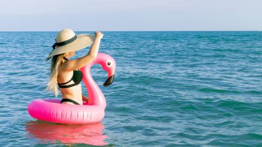 Yaz kumsalı dinleniyor. Bikini mayo, güneş gözlüğü ve pembe şişirilebilir flamingolu hasır şapkalı okyanus arka planında mavi deniz suyunda mutlu genç seksi kız. Moda yaz konsepti