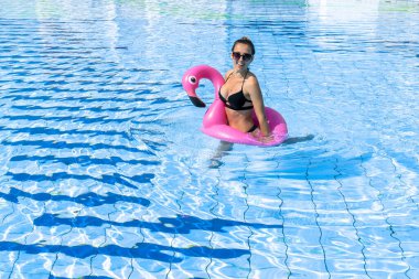 Bilardodaki kız. Bikini mayo giymiş, pembe şişirilebilir flamingolu güneş gözlüklü, mavi suda dinlenen mutlu ve seksi bir kadın. Yaz tatili eğlence özgürlüğü kavramı