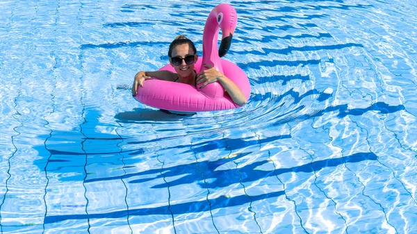 Summer pool relaxing. Young sexy woman in bikini swimsuit, sunglasses float with pink flamingo in blue water. Trendy summer concept