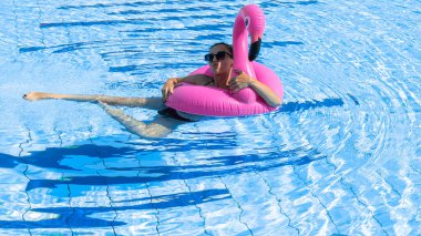 Bilardodaki kız. Bikini mayo giymiş, pembe şişirilebilir flamingolu güneş gözlüklü, mavi suda dinlenen mutlu ve seksi bir kadın. Yaz tatili eğlence özgürlüğü kavramı