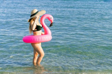 Tatil köyü refahı. Bikini mayo, güneş gözlüğü ve pembe şişirilebilir flamingolu hasır şapkalı okyanus kıyısı arka planında mavi deniz suyunda mutlu genç seksi kız. Tatil tatilinde..