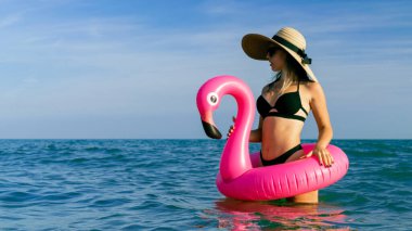 Plaj partisi kızı. Bikini mayo, güneş gözlüğü ve pembe şişirilebilir flamingolu hasır şapkalı okyanus sahilindeki mavi deniz suyunda mutlu genç seksi kız. Tatil tatili için otelde spa.