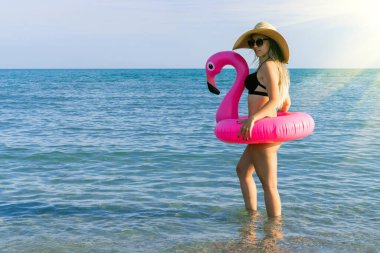 Merhaba yaz. Hasır şapkalı genç seksi kadın, bikini mayo, deniz sahili arka planı için mavi okyanus suyunda pembe şişirilebilir flamingo ile güneş gözlüğü. Moda yaz konsepti