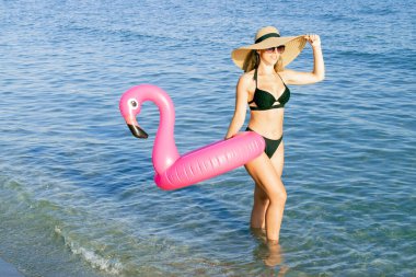 Yaz eğlencesi. Hasır şapkalı genç seksi kadın, bikini mayo, deniz sahili arka planı için mavi okyanus suyunda pembe şişirilebilir flamingo ile güneş gözlüğü. Kaplıca ve güneşlenme açık havada