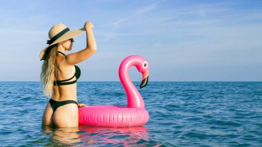 Yaz şapkası plajı. Bikini mayo, güneş gözlüğü ve pembe şişirilebilir flamingolu hasır şapkalı okyanus arka planında mavi deniz suyunda mutlu genç seksi kız. Lüks yaşam tarzı seyahati