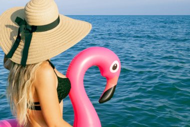 Yüzün genç bayan. Bikini mayo, güneş gözlüğü ve pembe şişirilebilir flamingolu hasır şapkalı okyanus kıyısı arka planında mavi deniz suyunda mutlu genç seksi kız. Lüks yaşam tarzı seyahati