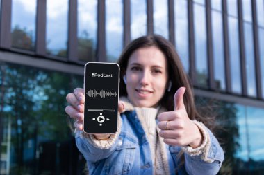 Podcast arka planı. Mobil akıllı telefon ekranlı, podcast uygulamalı, sesli kulaklıklı mutlu genç seksi kız. Ses telsiz cihazları kadının elinde. Kayıt stüdyosu arka planı