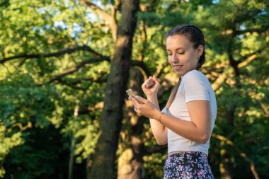 Gülümseyen kız cep telefonu. Parktaki güneşli bir günde elinde akıllı telefon tutan neşeli genç bir kız. Mesajlaşma, mesaj atmaktan mutlu olmak.