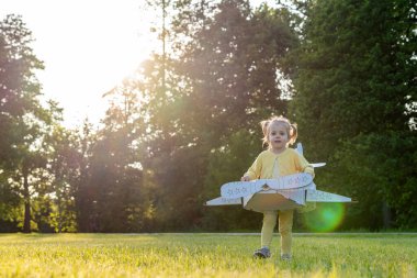 Karton uçak. Çocuk pilot dışarıda oynuyor. Bebek kostümlü kız tarlası sarısı