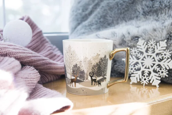 Geyiklerin içinde altın ve beyaz kış fincanı. Pencere kenarında çay ya da kahve, süveterin yanında, kış süslemesi.