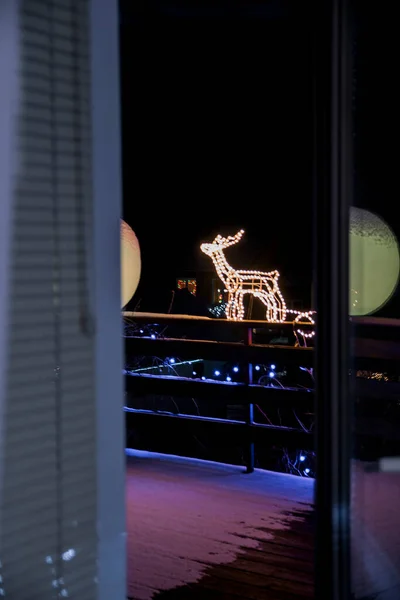 Karla kaplı balkonun penceresinden Noel ışıklarıyla parlayan bir ren geyiği ve kızak.