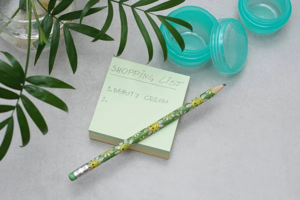 A Post-it Yapışkan Notlar 'da el yazısı bir alışveriş listesi. Cilt kremi için boş markasız kavanozlar. Gri arka planda etiketsiz yeşil plastik kaplar..