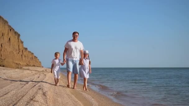 若い白人の家族がビーチを散歩しています。父の息子と娘は砂の上で手を握って歩く — ストック動画