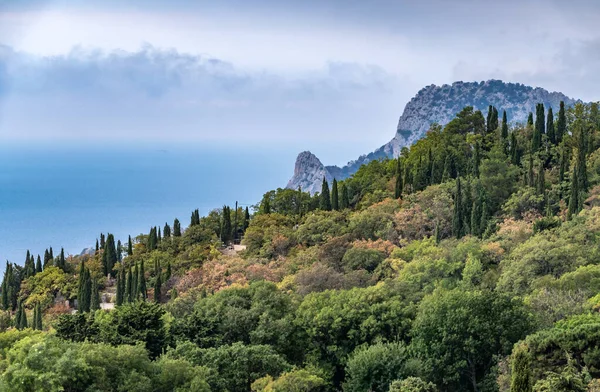 Rocky mountain on sea shore with high green trees on slope. Seascape with cloudy sky. Old cemetery on sea shore