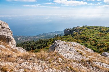 Günbatımında konut binaları olan yeşil deniz kıyısı. Yamaçta yüksek yeşil ağaçlarla deniz kıyısında kayalık bir dağ