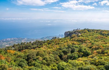 Günbatımında konut binaları olan yeşil deniz kıyısı. Yamaçta yüksek yeşil ağaçlarla deniz kıyısında kayalık bir dağ